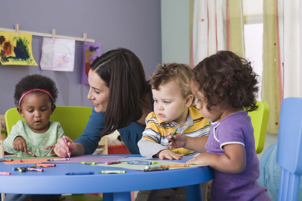 Picture of educator showing three toddlers how to use crayons